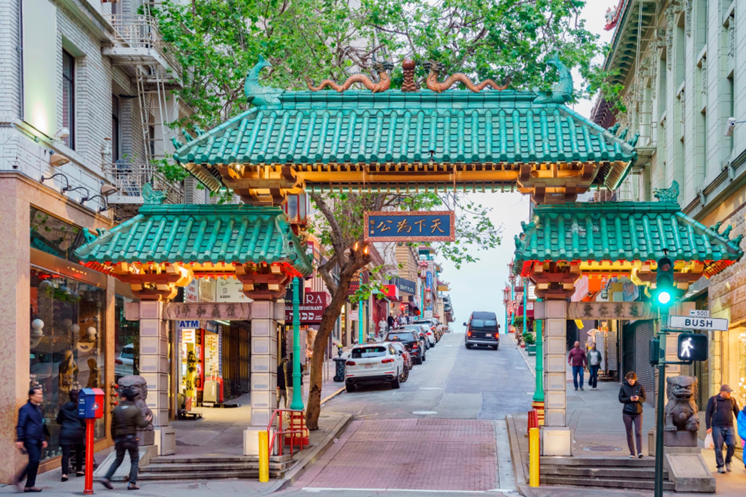 Ornate Chinese gate spans a bustling city street, surrounded by shops and pedestrians, with vehicles parked along the road. Traffic lights indicate crossing.