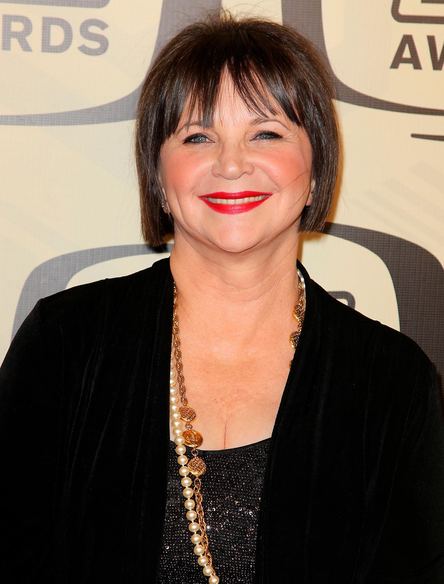 Woman smiling, wearing a black outfit with a pearl necklace, standing against a backdrop with the word "AWARDS."