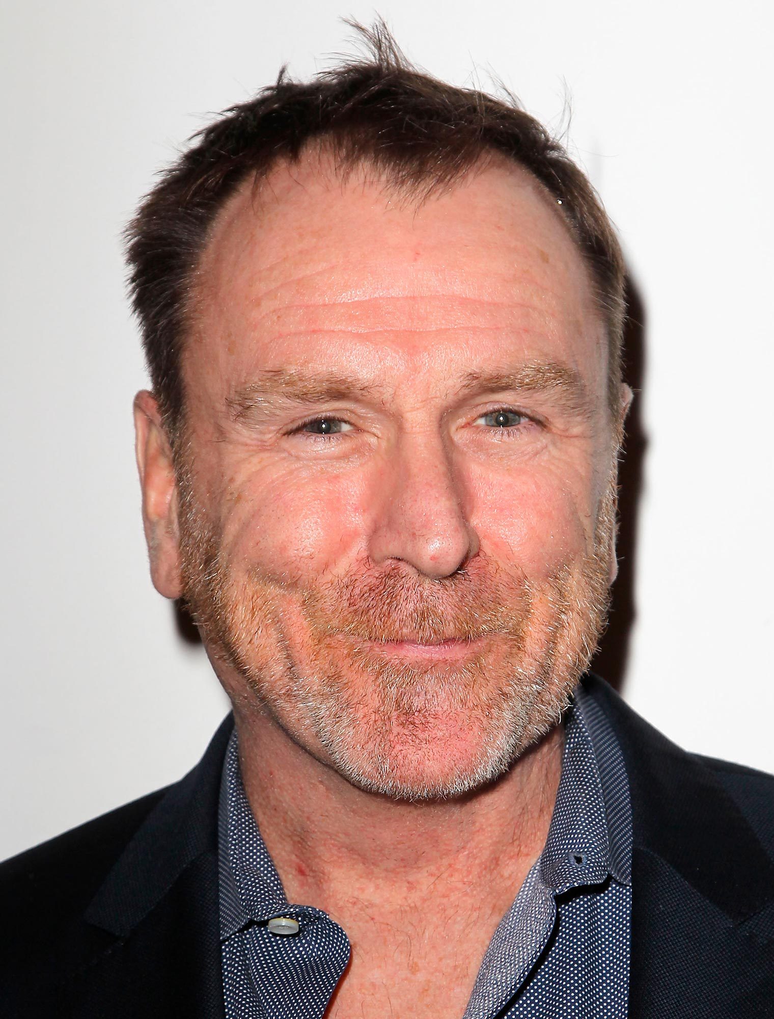 A man is smiling, wearing a navy blazer and dotted shirt against a plain, white background.