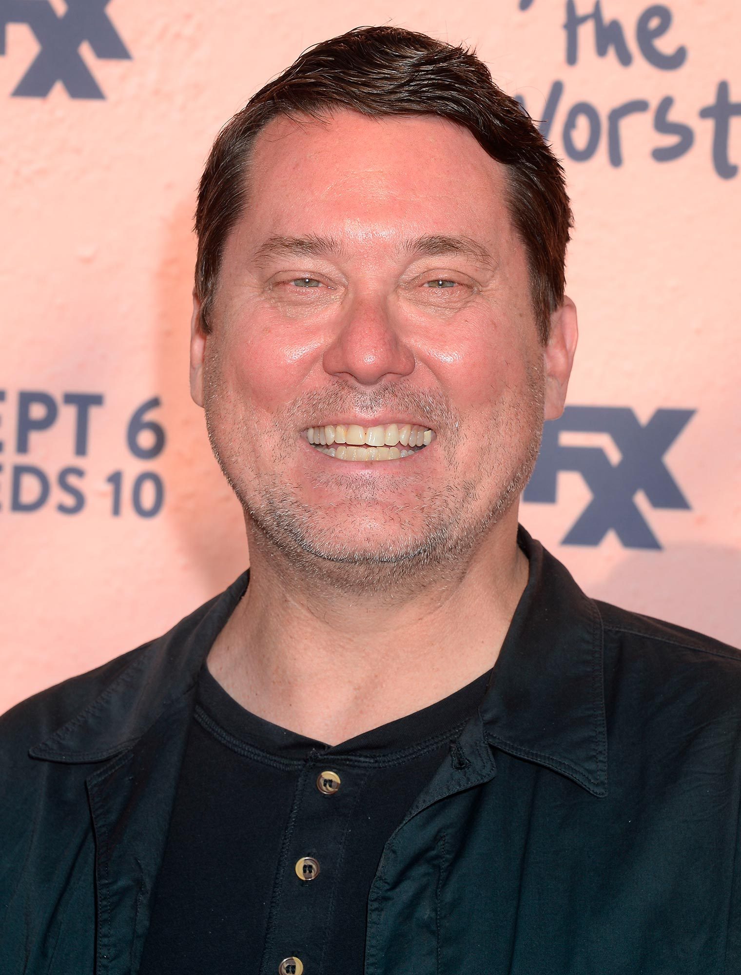 A person smiling in front of a peach-colored backdrop with the text â€œFXâ€ and â€œSEPT 6 WEDS 10â€ partially visible.