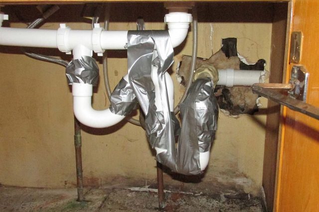 Pipes wrapped in duct tape, poorly secured, under a wooden kitchen cabinet with a hole in the wall behind them.