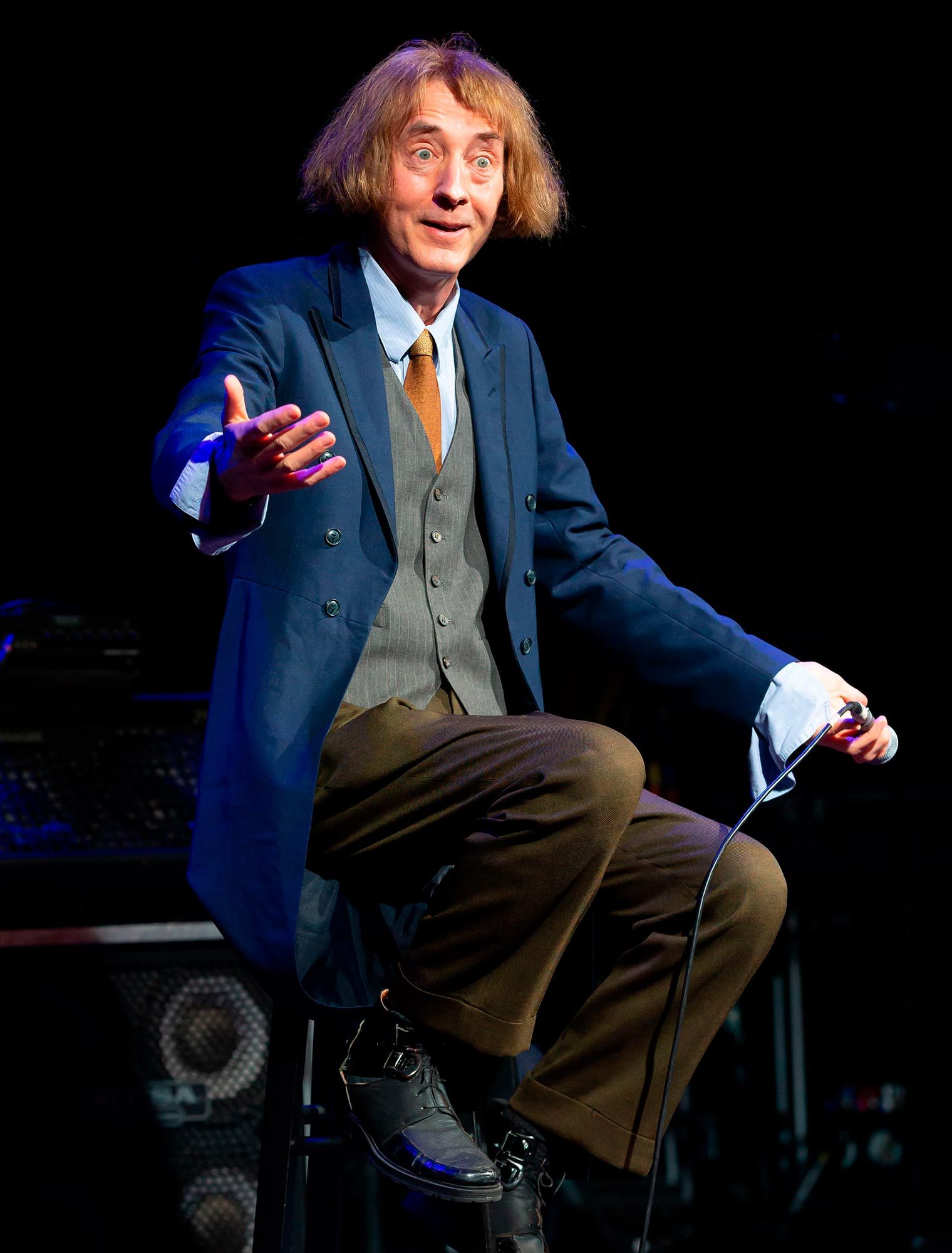 Performer gestures while sitting, holding a microphone on stage, wearing a blue jacket, gray vest, and brown trousers, against a dark background.