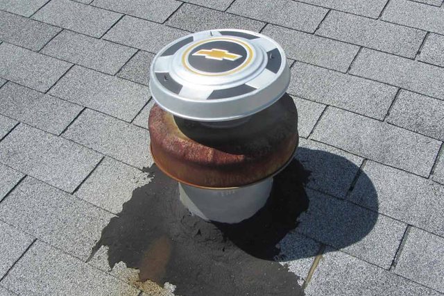 Hubcap covers a roof vent pipe, surrounded by asphalt patch on shingled roof.