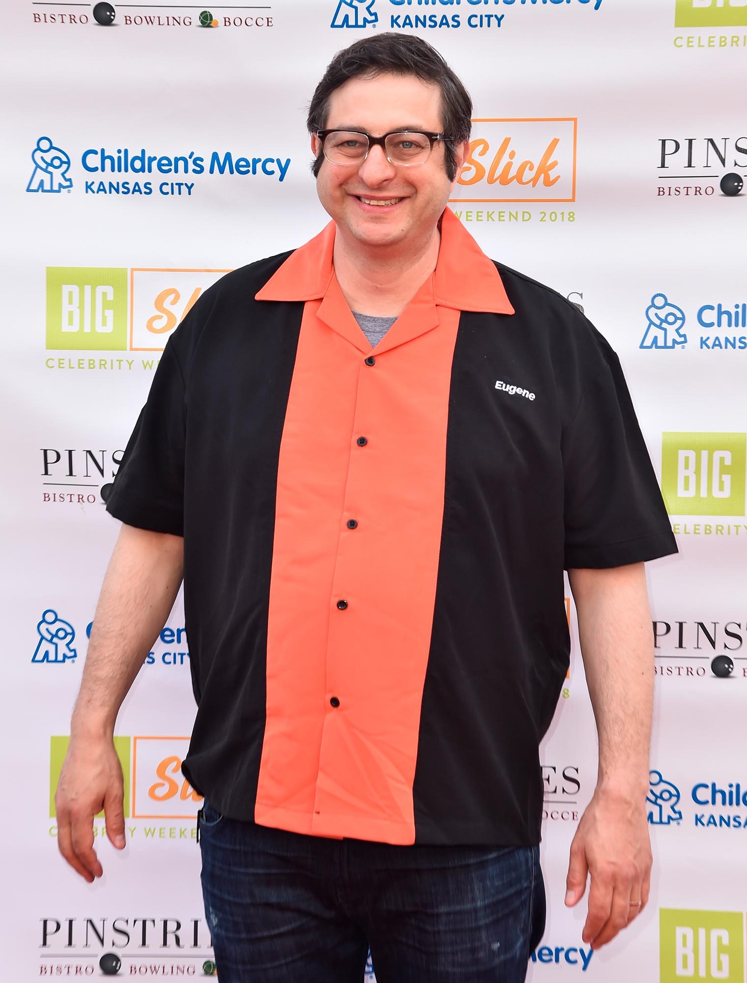 A person wearing a black and orange shirt smiles against a backdrop featuring various logos, including "Children's Mercy Kansas City" and "Slick."