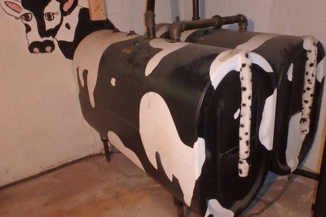 Oil tank painted like a cow, standing in an unfinished basement, with pipe fittings resembling horns and a cow face on the wall.