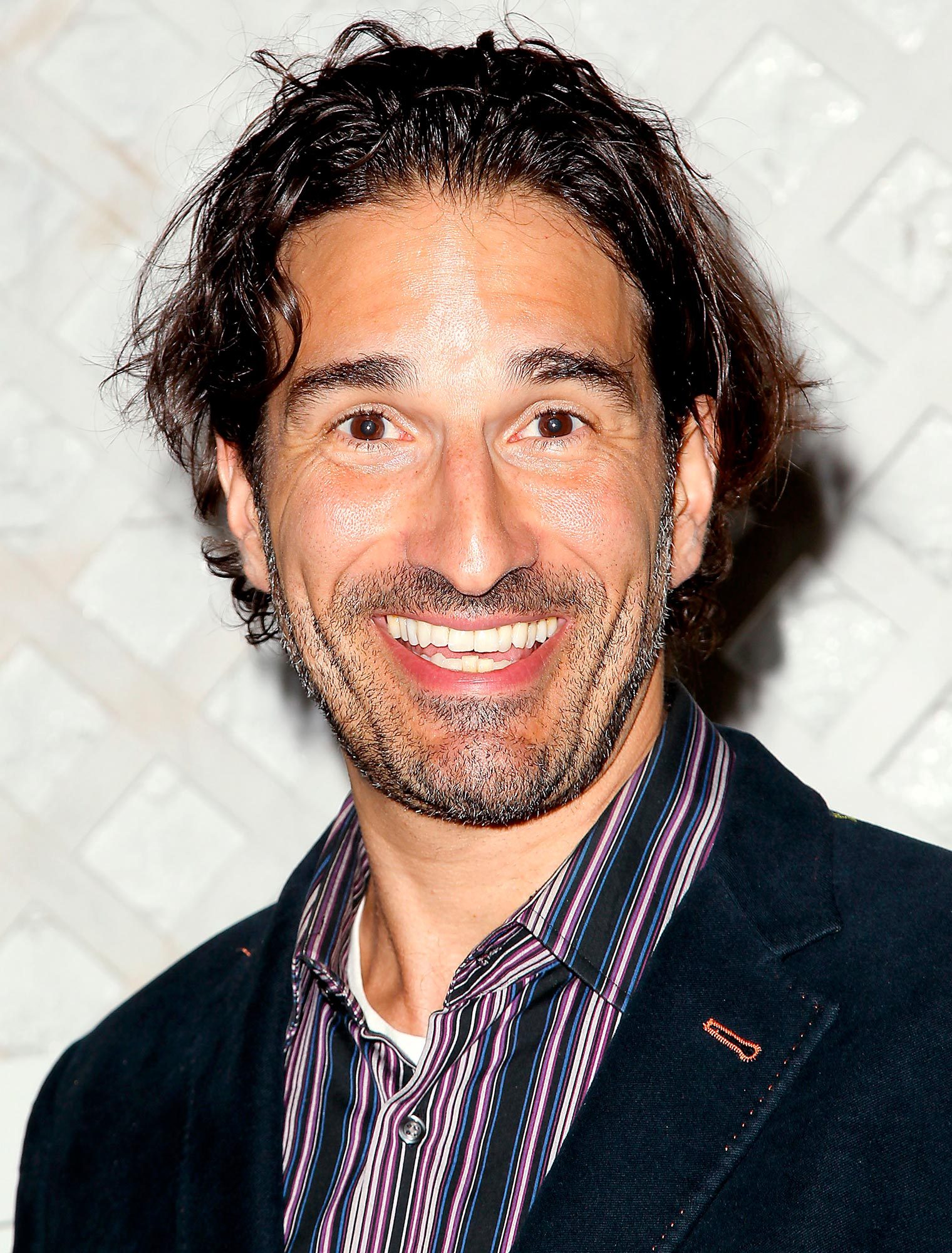 A smiling man faces forward, wearing a striped shirt and dark blazer, with a textured light background.