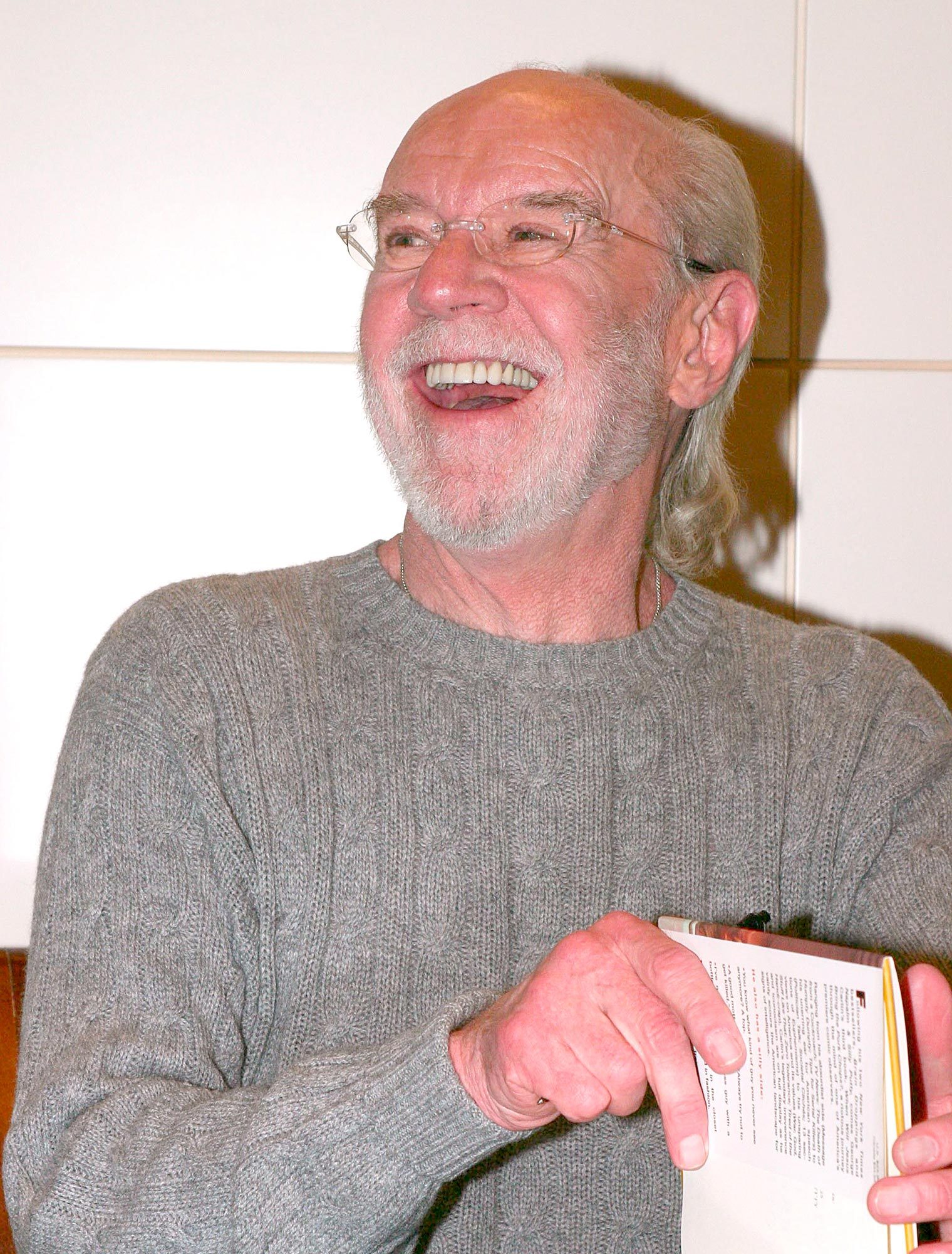A bearded man, smiling broadly, holds a book in a light-colored room.