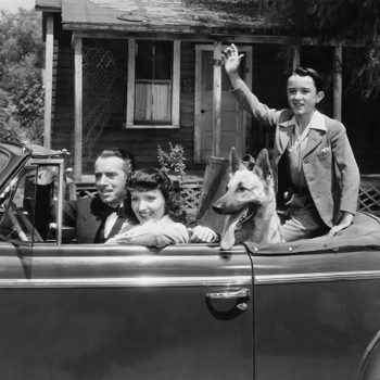 A family sits in a convertible car. A man drives, a woman smiles, a boy waves, and a dog sits beside them. Rural house background.