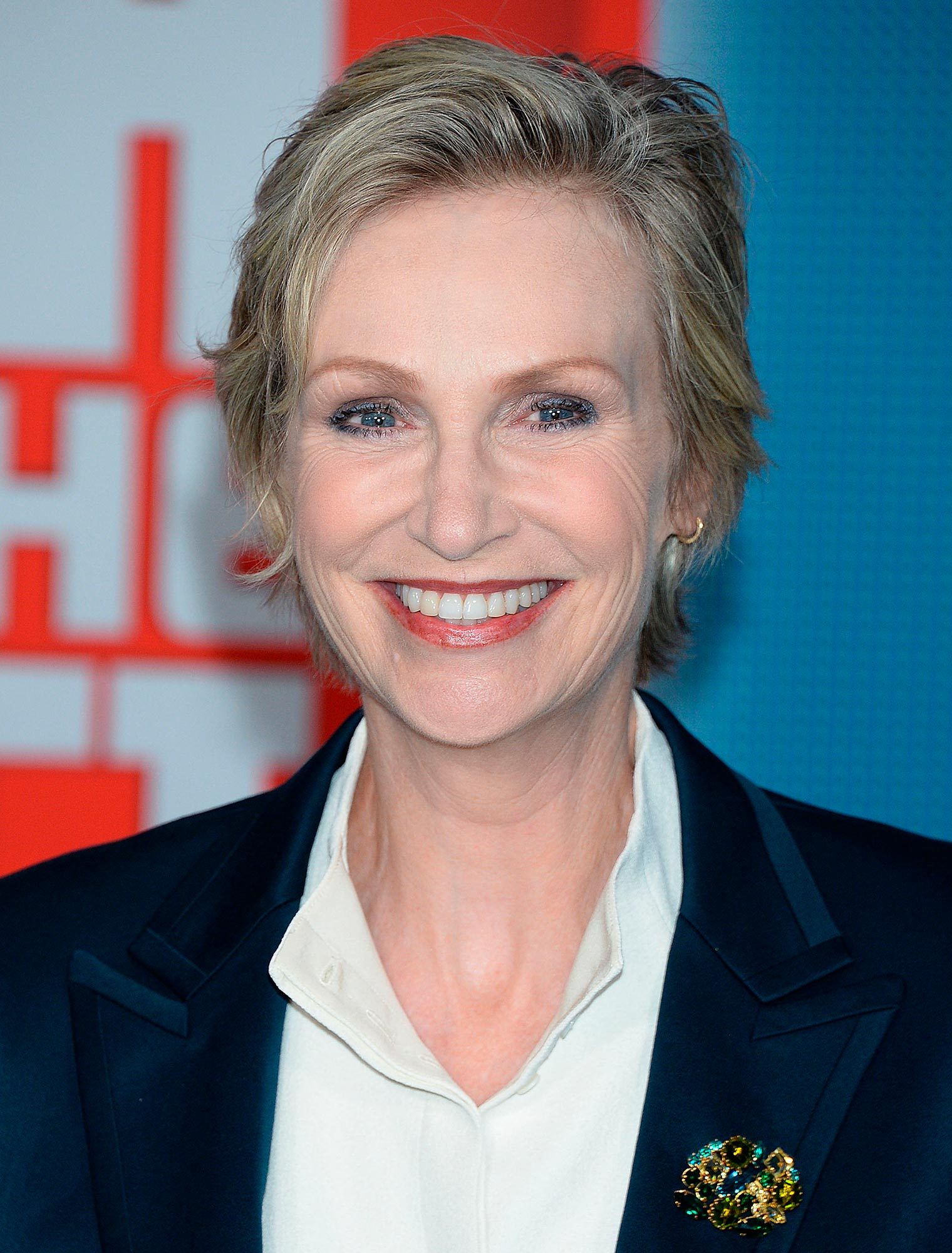 Smiling person wearing a dark blazer and white shirt, standing against a colorful background with large, indistinct text.