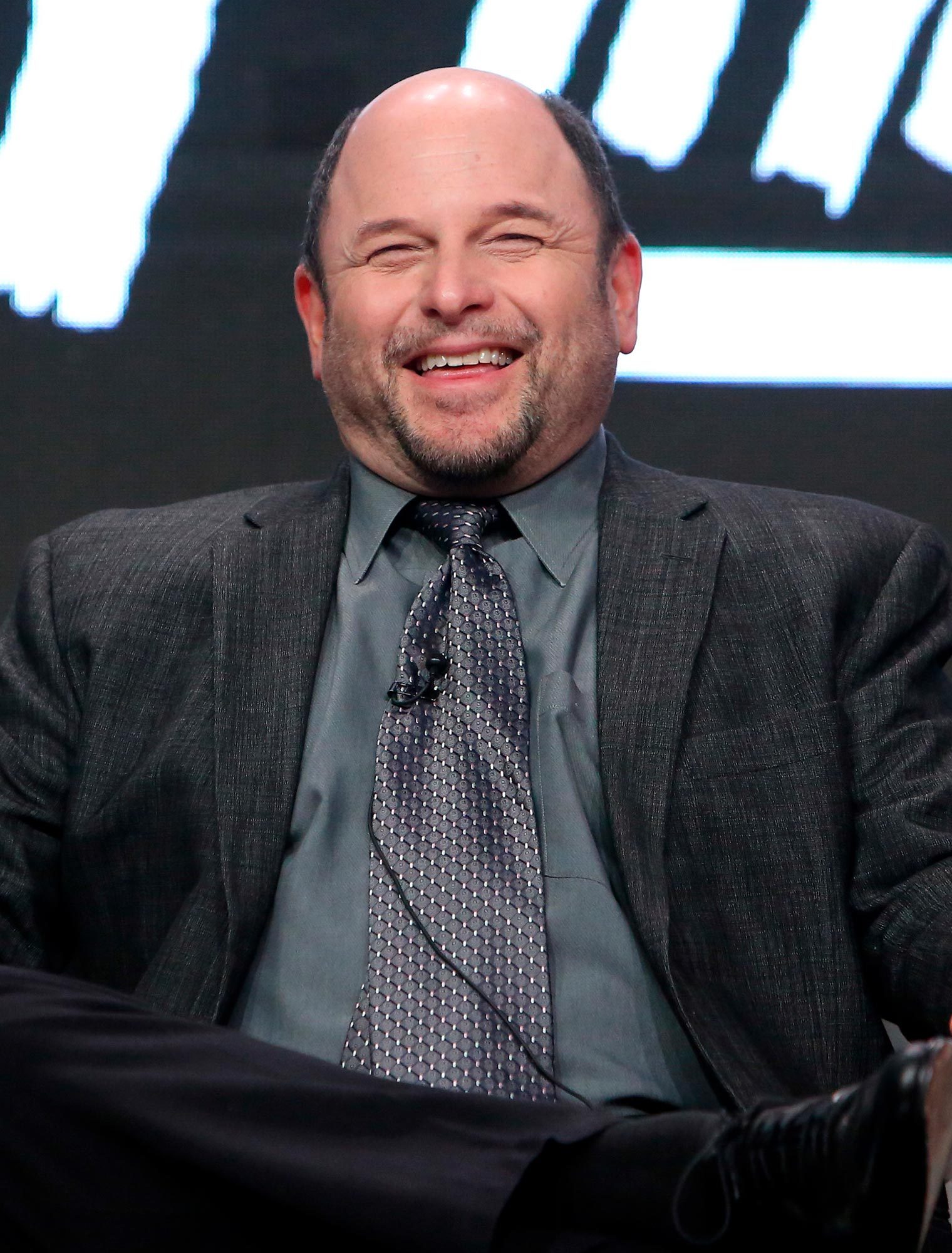 A man wearing a suit is smiling broadly, seated onstage against a dark, blurred background.