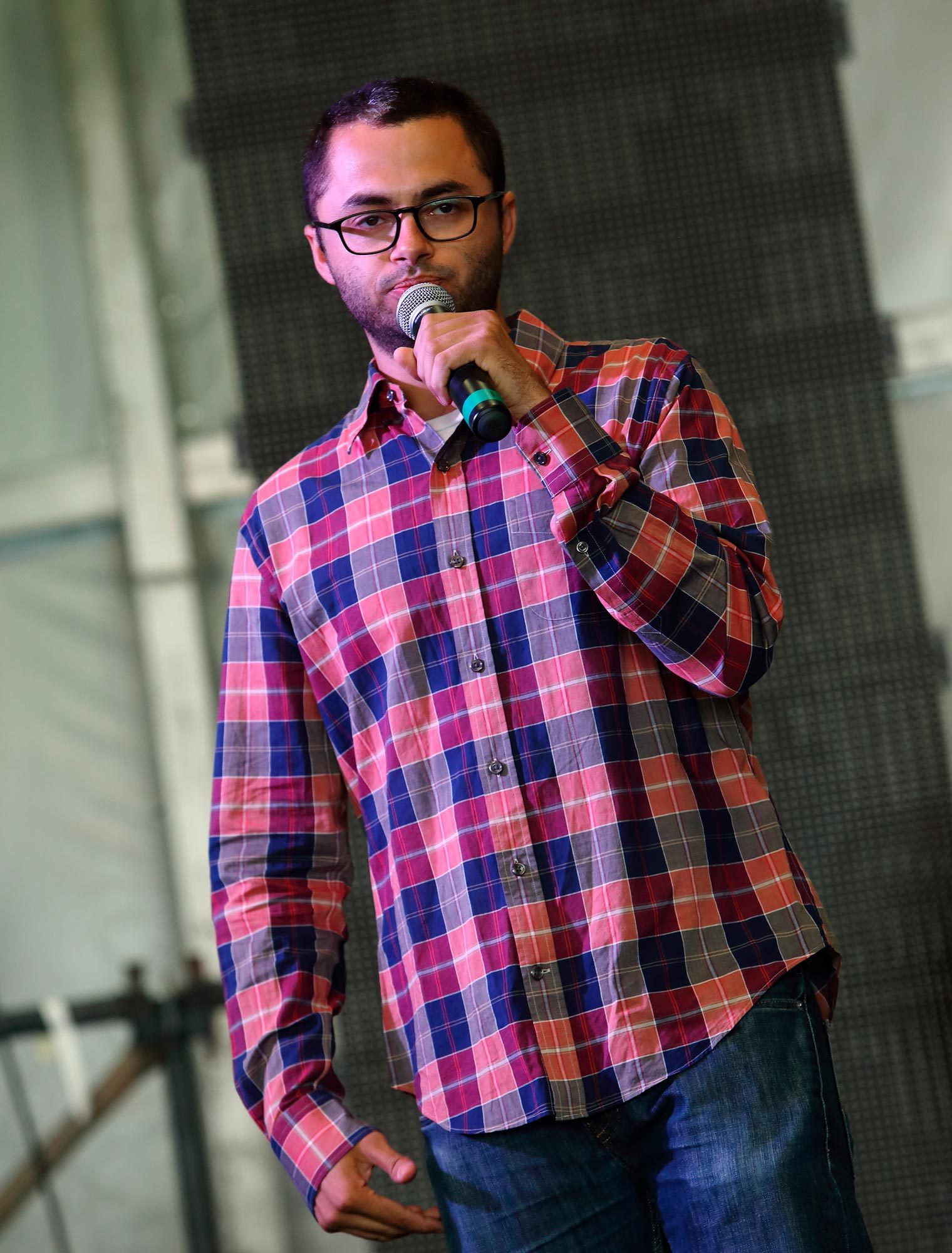 A person wearing glasses and a plaid shirt speaks into a microphone on a stage with a blurred background.