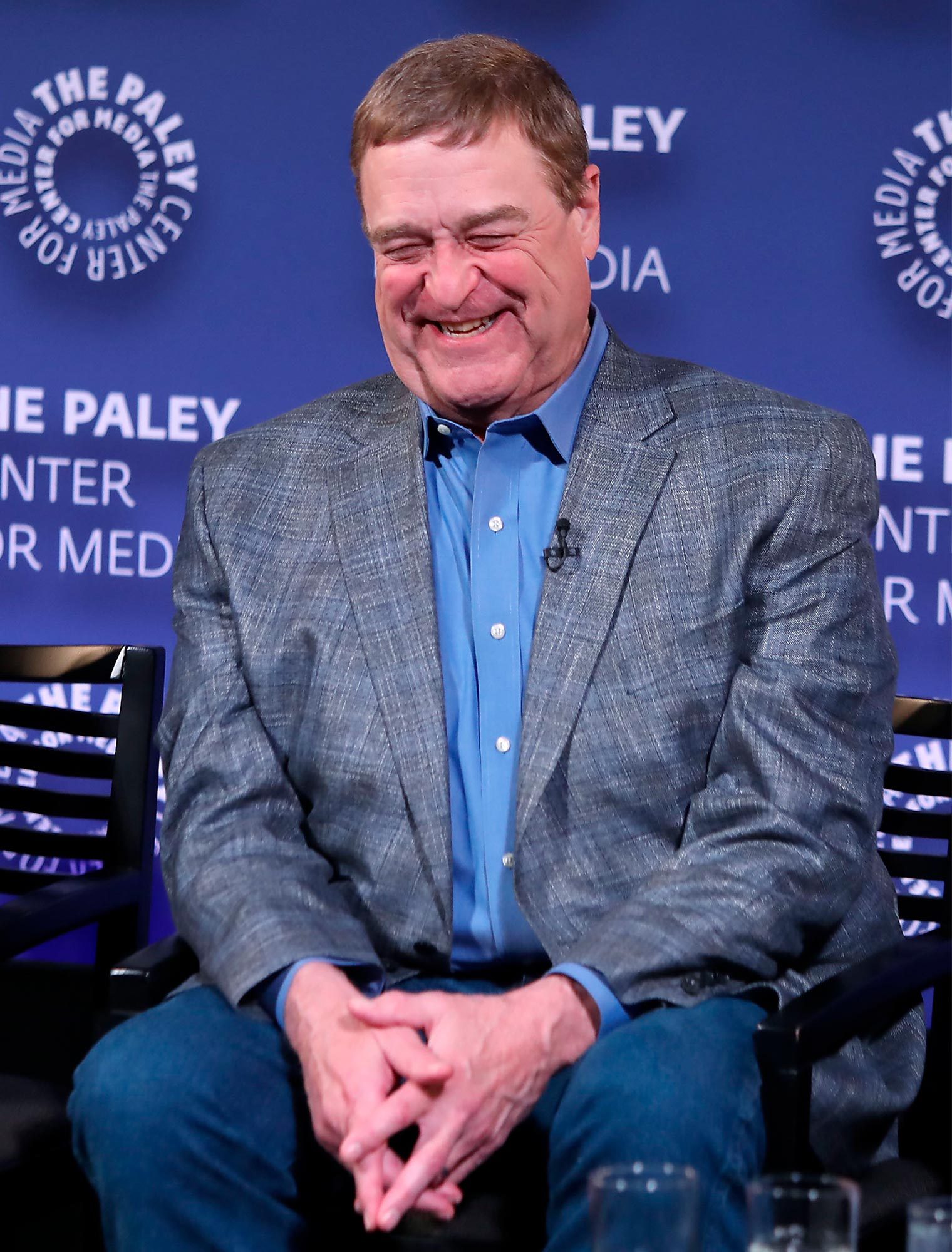 A person smiles while sitting on a chair, wearing a gray blazer. The background displays "The Paley Center for Media" in blue.