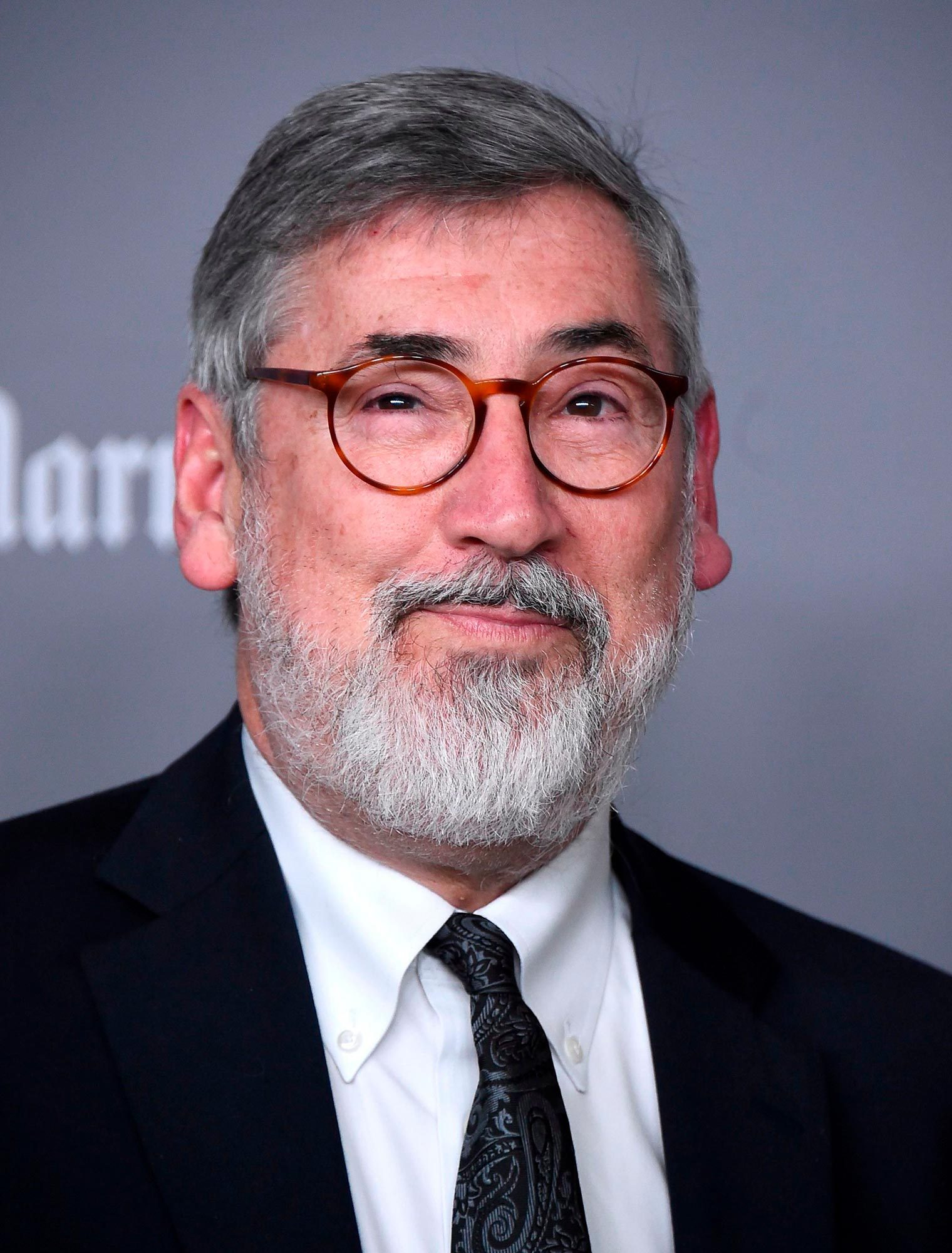 Man with gray hair and beard, wearing glasses and a suit, poses against a plain gray background, slightly smiling.
