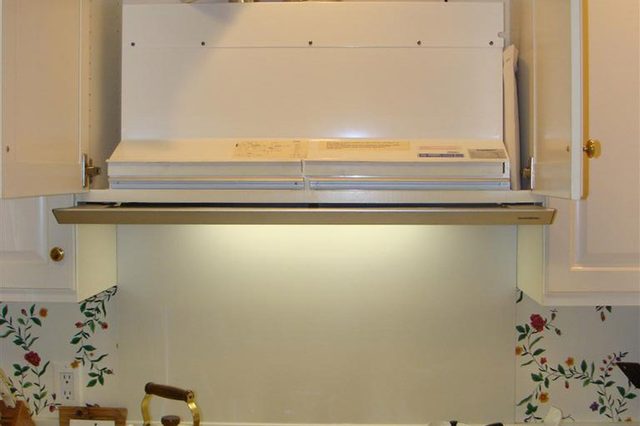 Stovetop displays teapots and utensils, beneath a lit range hood with exposed ductwork, in a kitchen adorned with floral wallpaper and white cabinets.