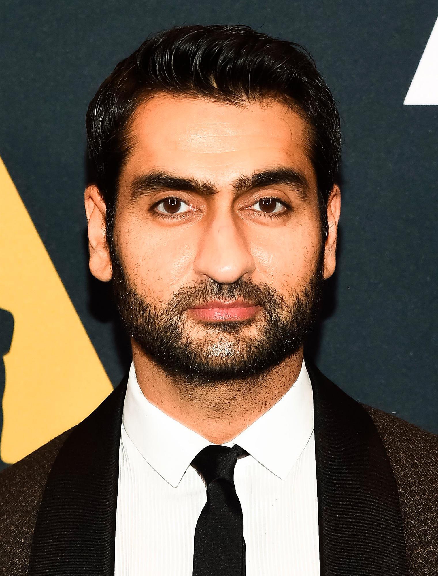 A man, in formal attire, stands against a dark backdrop. He wears a white shirt, black tie, and dark jacket.