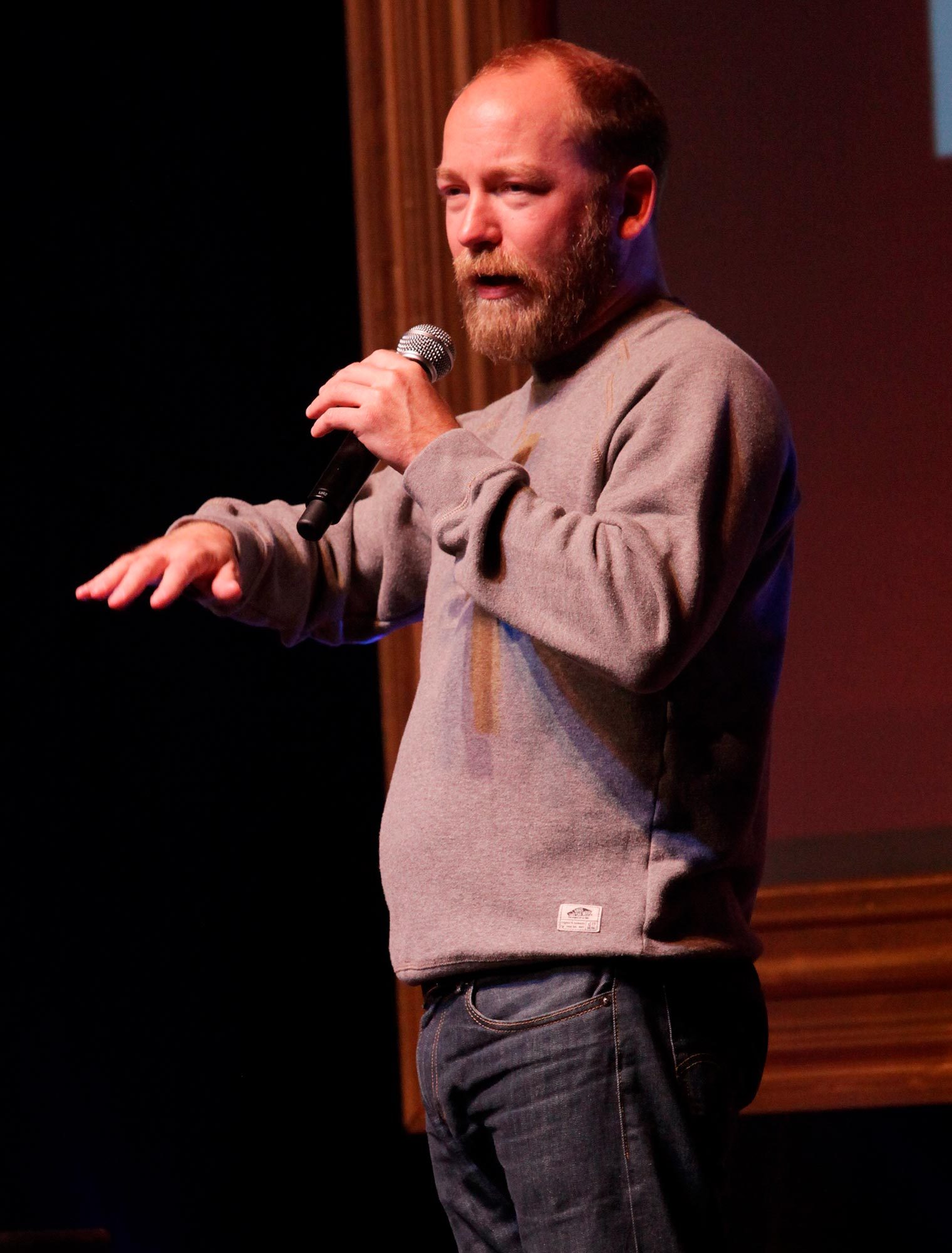Man speaks into a microphone, gesturing with his hand, on a dimly lit stage with a dark background.