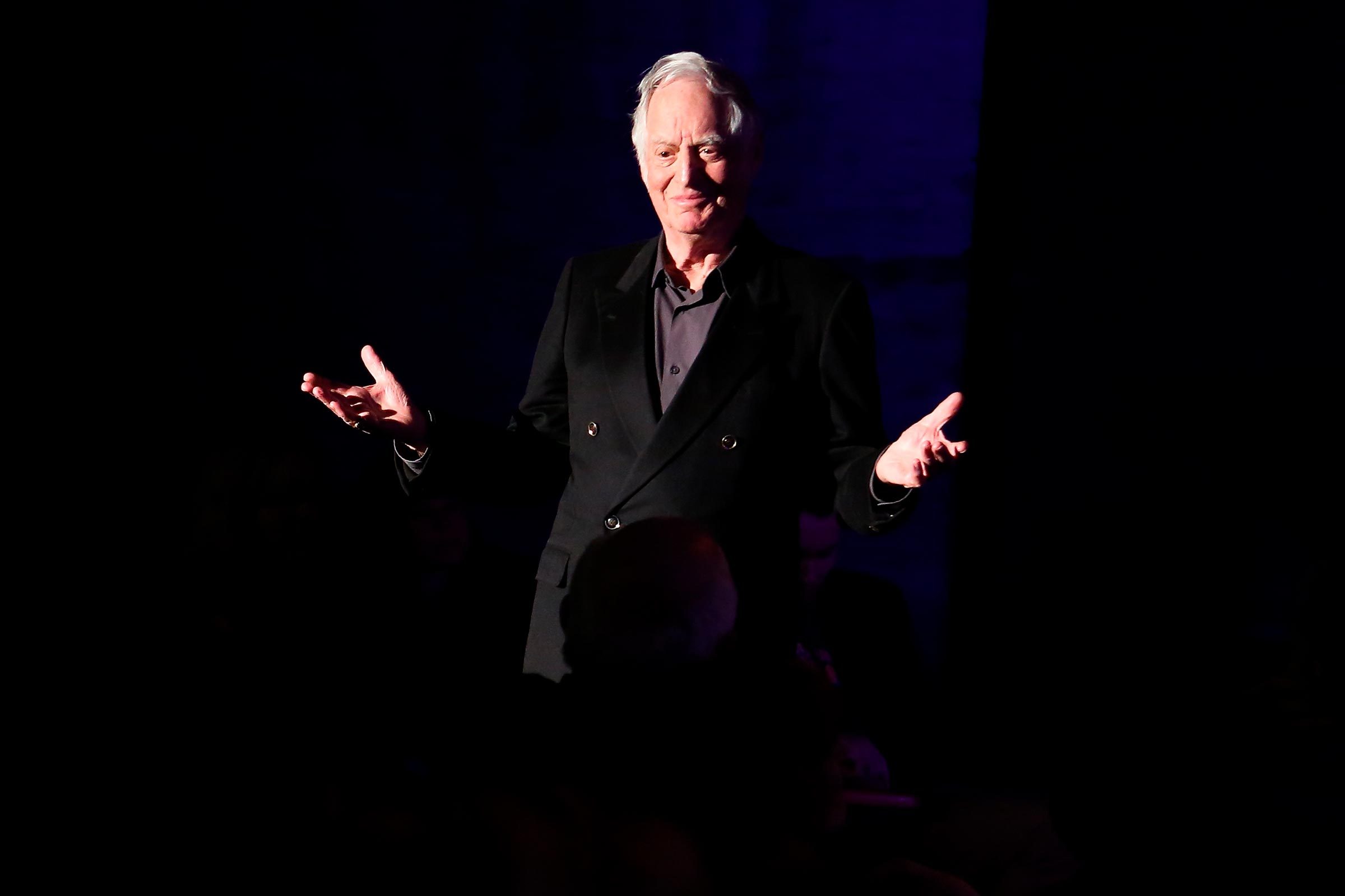Man stands gesturing with open hands, wearing a dark suit, on a dimly lit stage against a dark background.