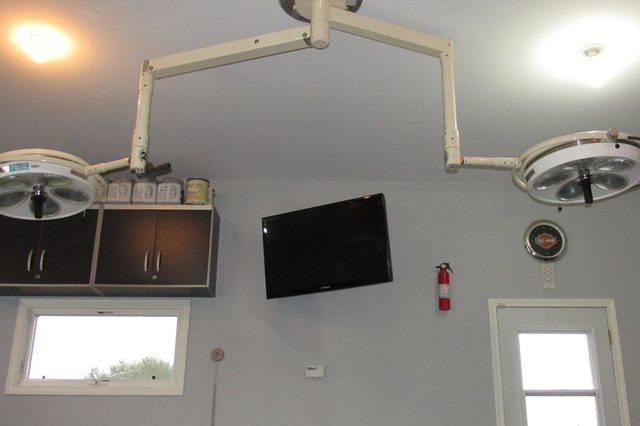 Ceiling-mounted surgical lights hang in a garage with a wall-mounted TV, fire extinguisher, clock, and a small window near the door.