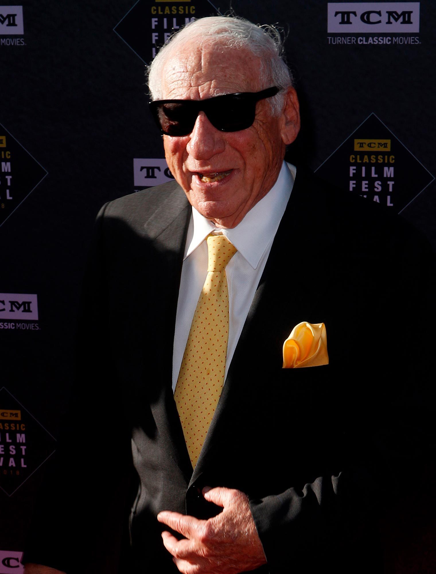 Man wearing sunglasses smiles, standing on a red carpet. He wears a black suit, yellow tie, and matching pocket square. Background features "TCM Classic Film Festival" logos.