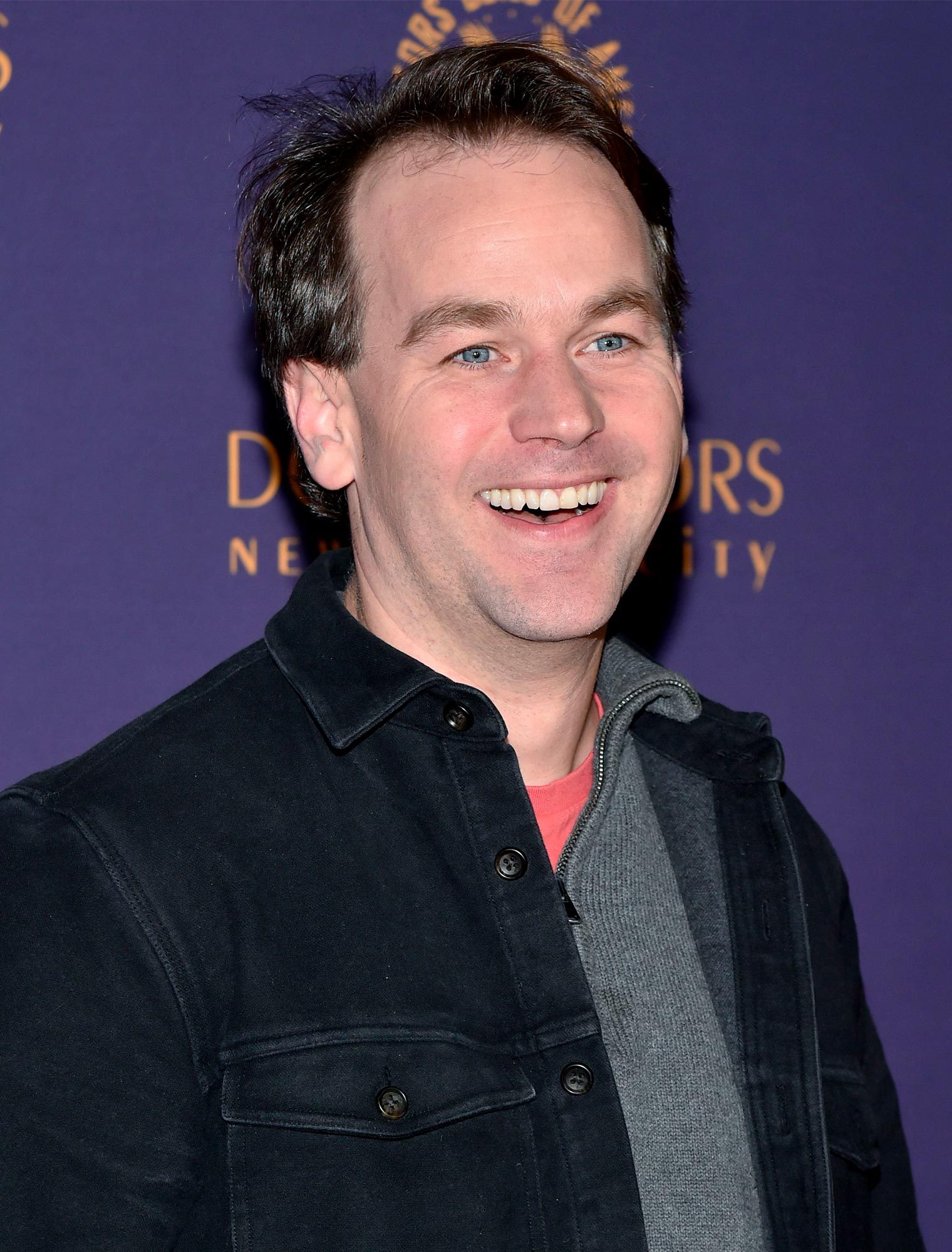 Person smiling, wearing a black jacket and grey sweater, standing in front of a purple backdrop with partially visible golden text.