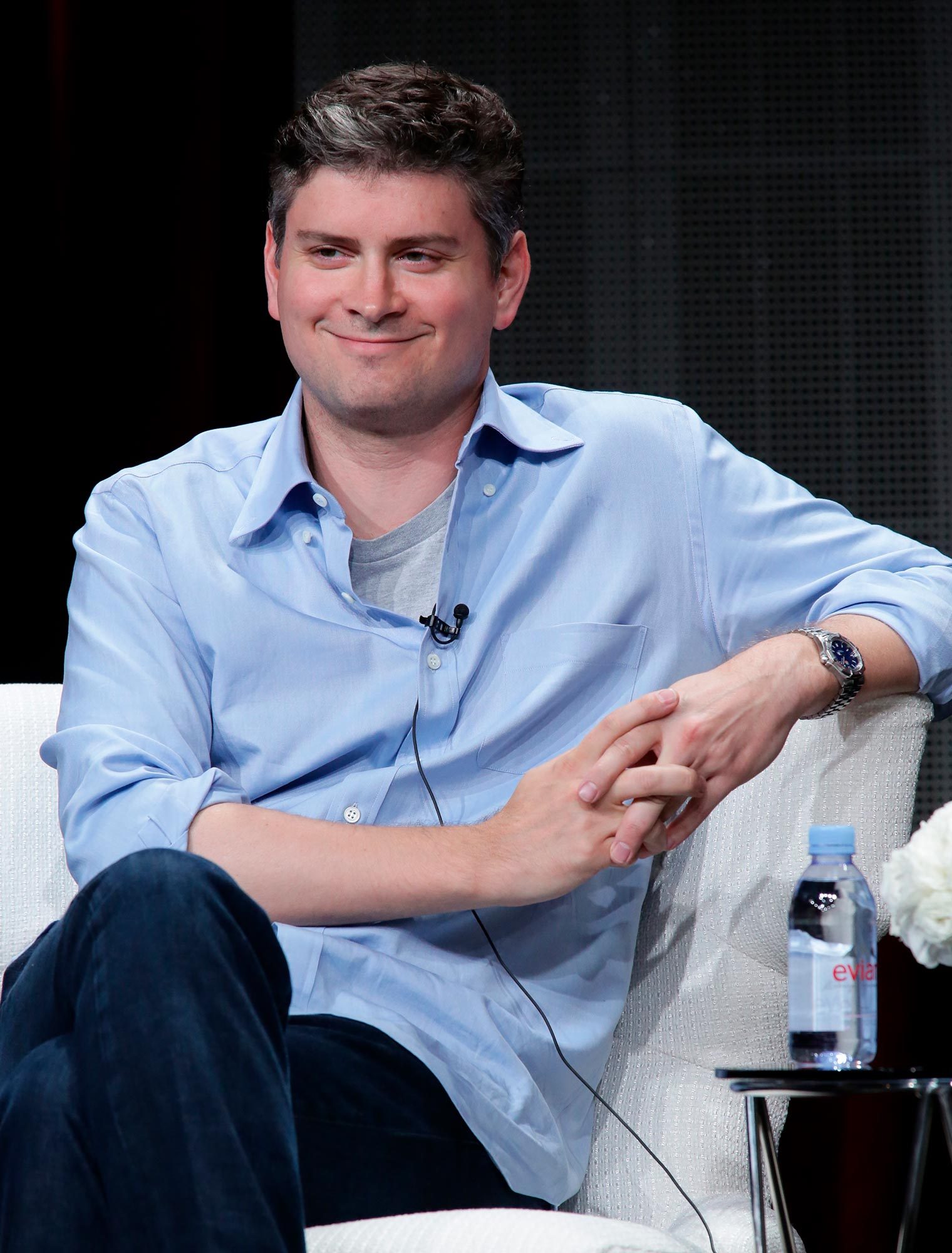 A person in a blue shirt smiles while sitting cross-legged on a white chair, holding hands. An Evian water bottle is nearby.