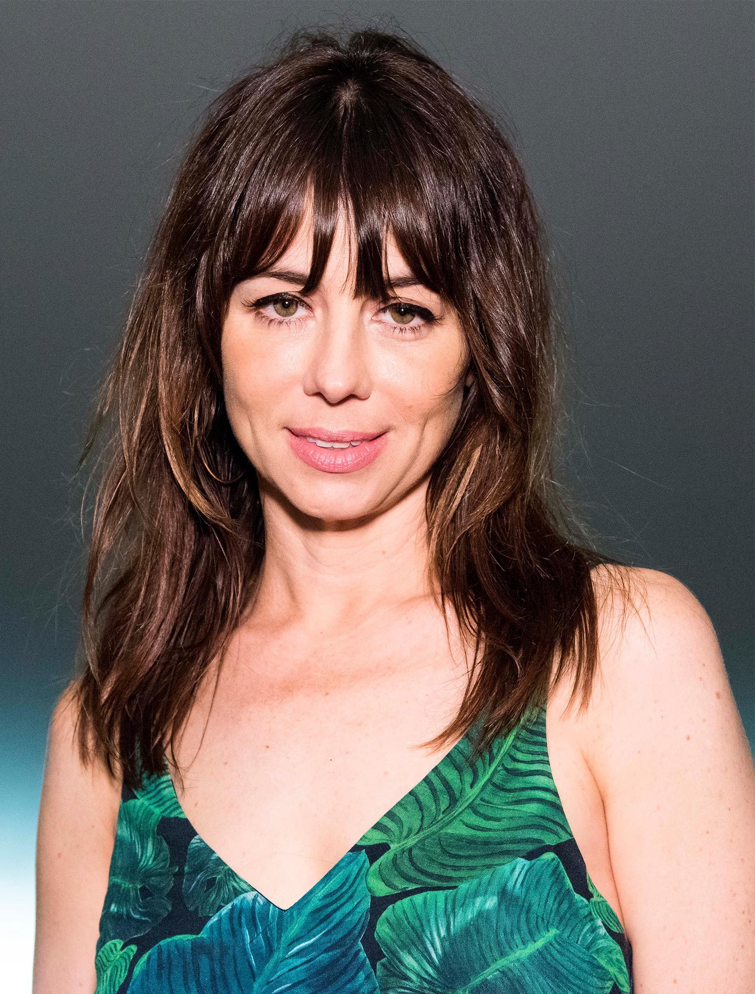 Woman smiling slightly, wearing a green leaf-patterned top, standing against a neutral gradient background.