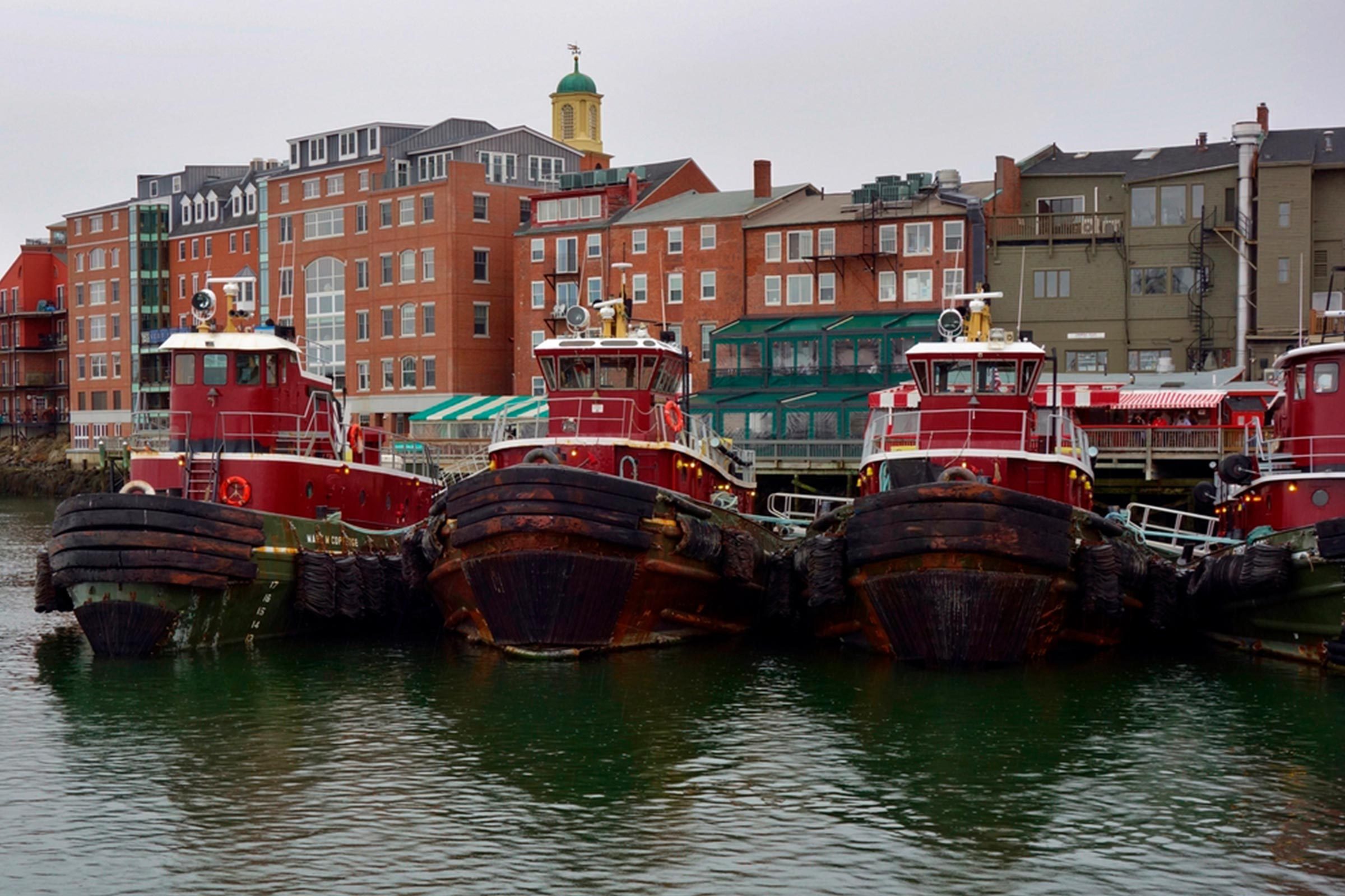 Tugboats docked together along a waterfront, bordered by several brick buildings under an overcast sky.