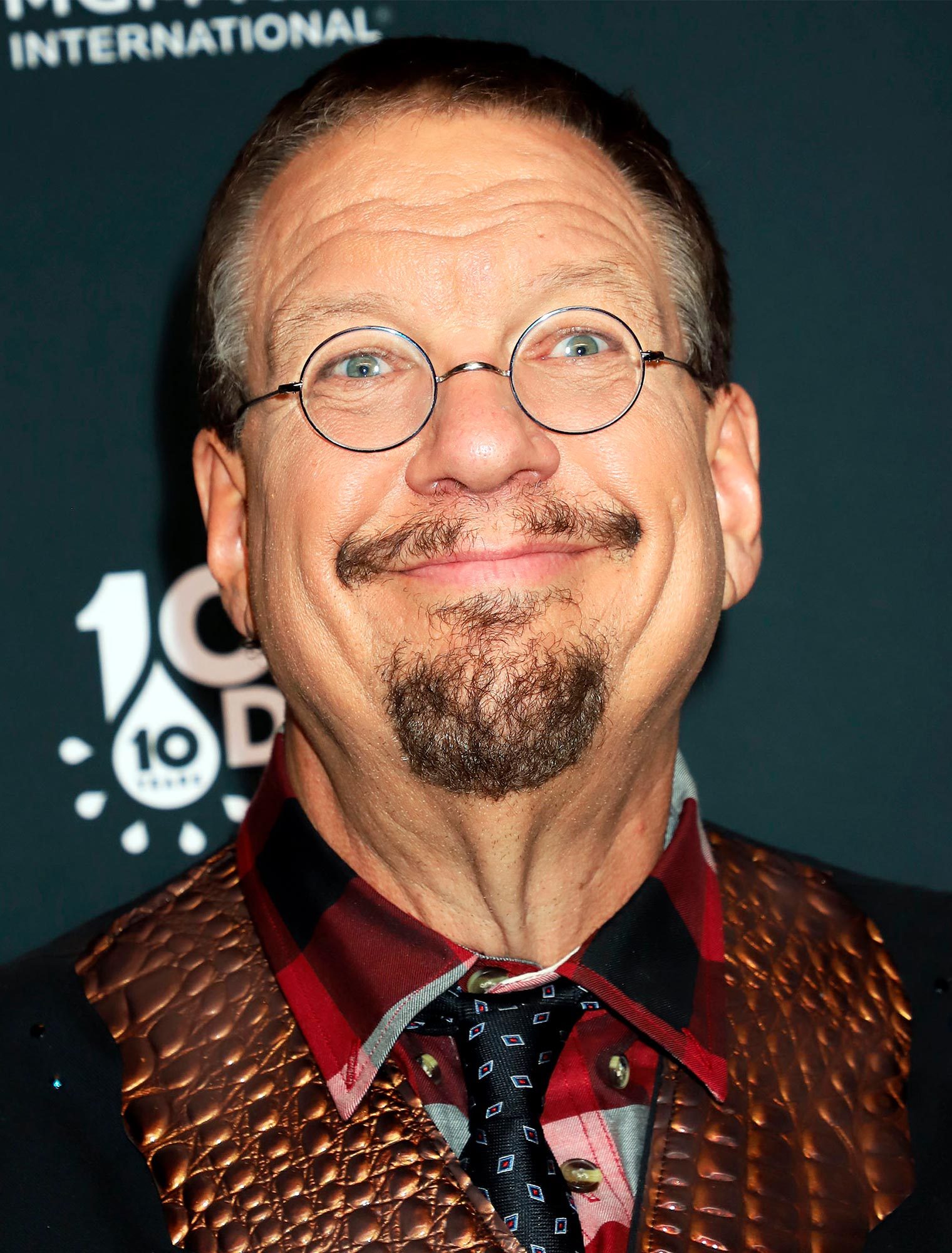 Man smiling broadly, wearing round glasses and a red plaid shirt with a textured vest, standing against a dark backdrop with partial text.