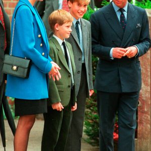 A smiling family stands outside, dressed formally. They appear to be engaged in conversation with each other, surrounded by a brick building and greenery.