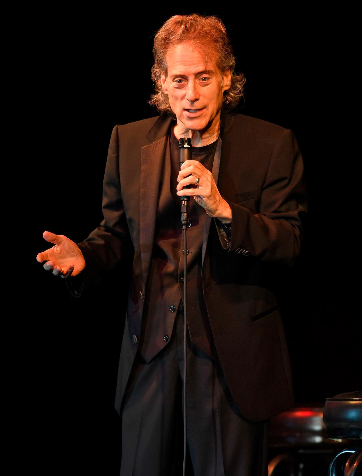 Man holds microphone, speaking animatedly on stage, wearing a black suit. Background is dark, with a hint of a stool beside him.