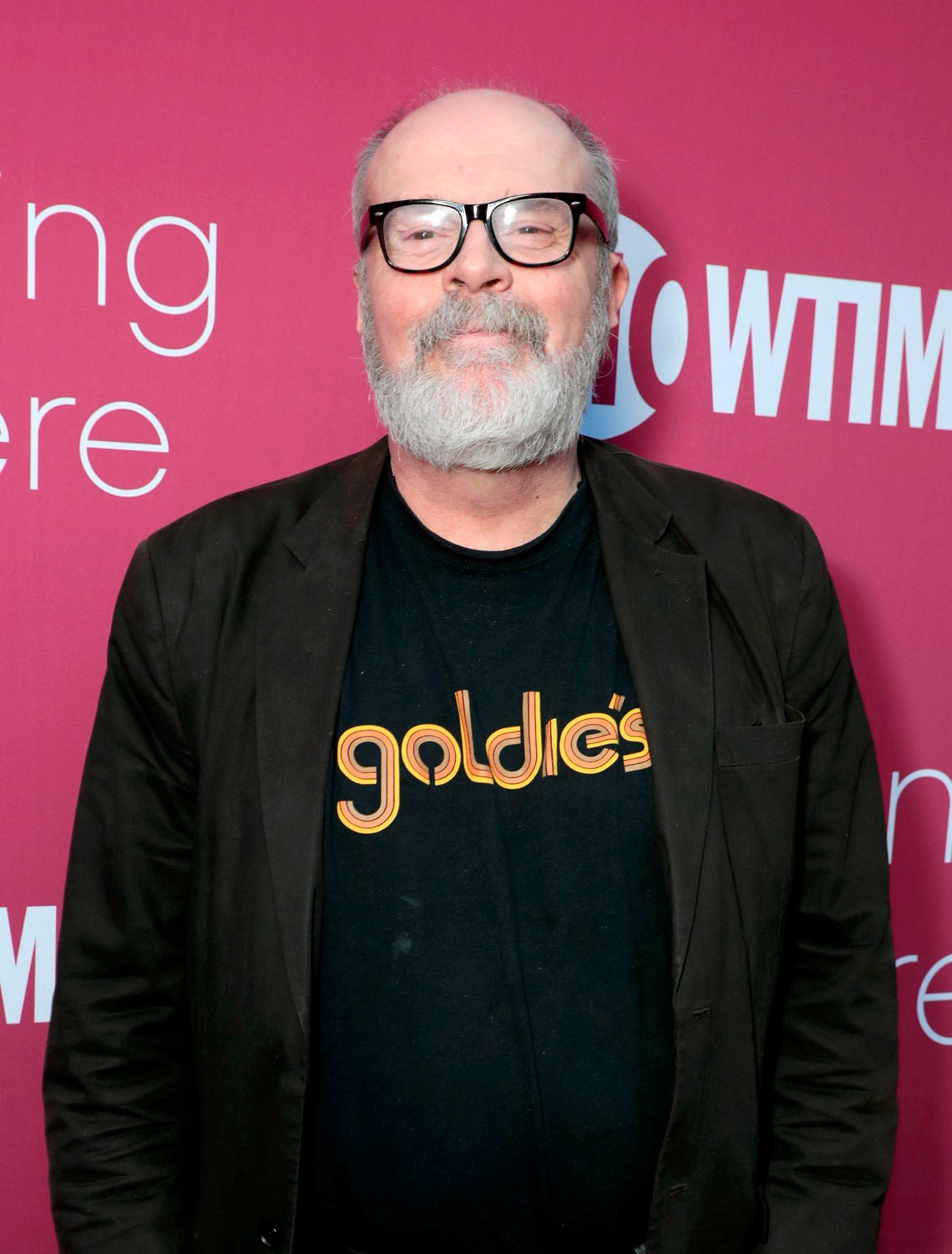 A person stands smiling, wearing glasses and a "goldie's" shirt, in front of a magenta backdrop with partially visible text "W TME".