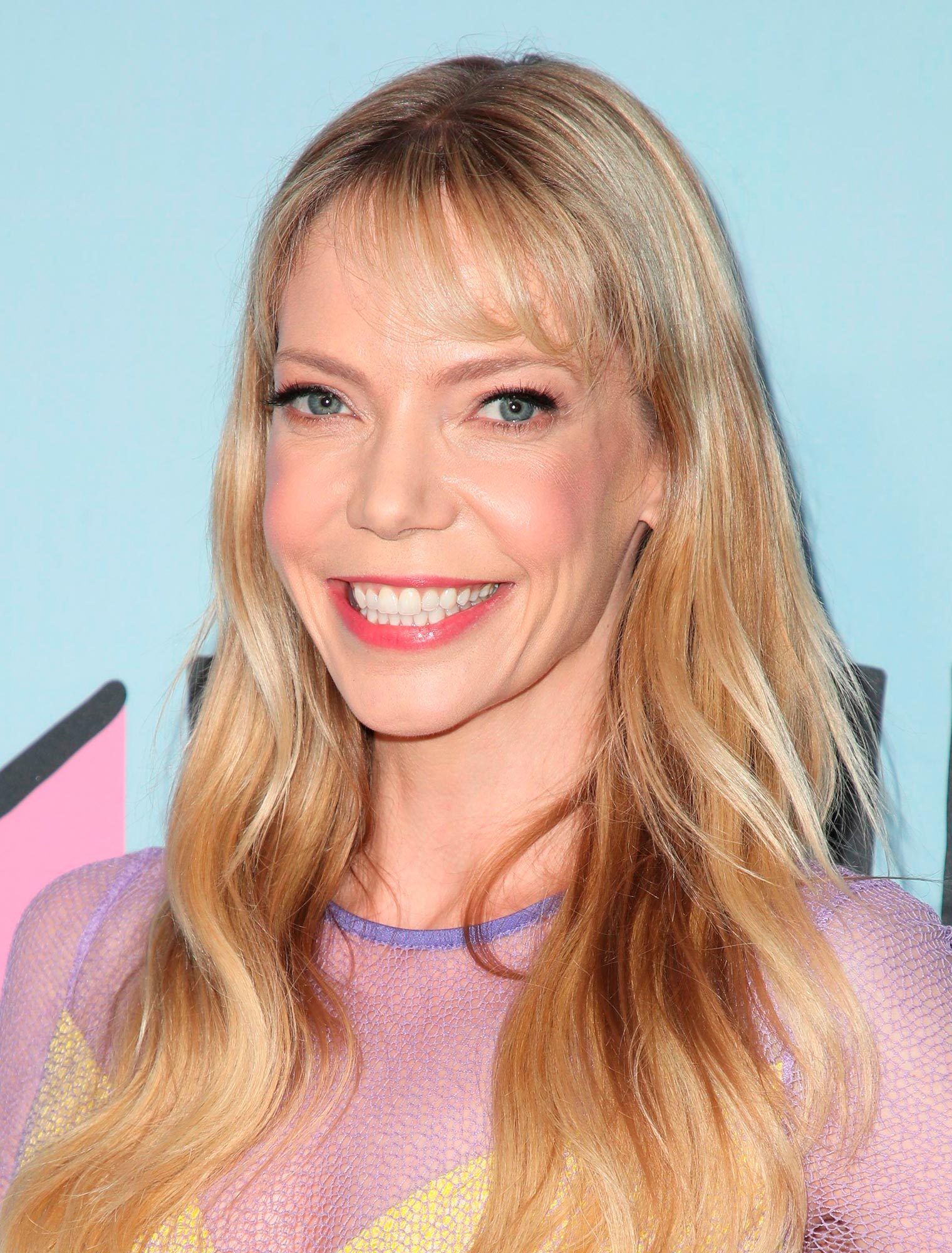 A person smiles broadly, with long blonde hair, wearing a colorful top. The background is a light blue wall with abstract shapes.
