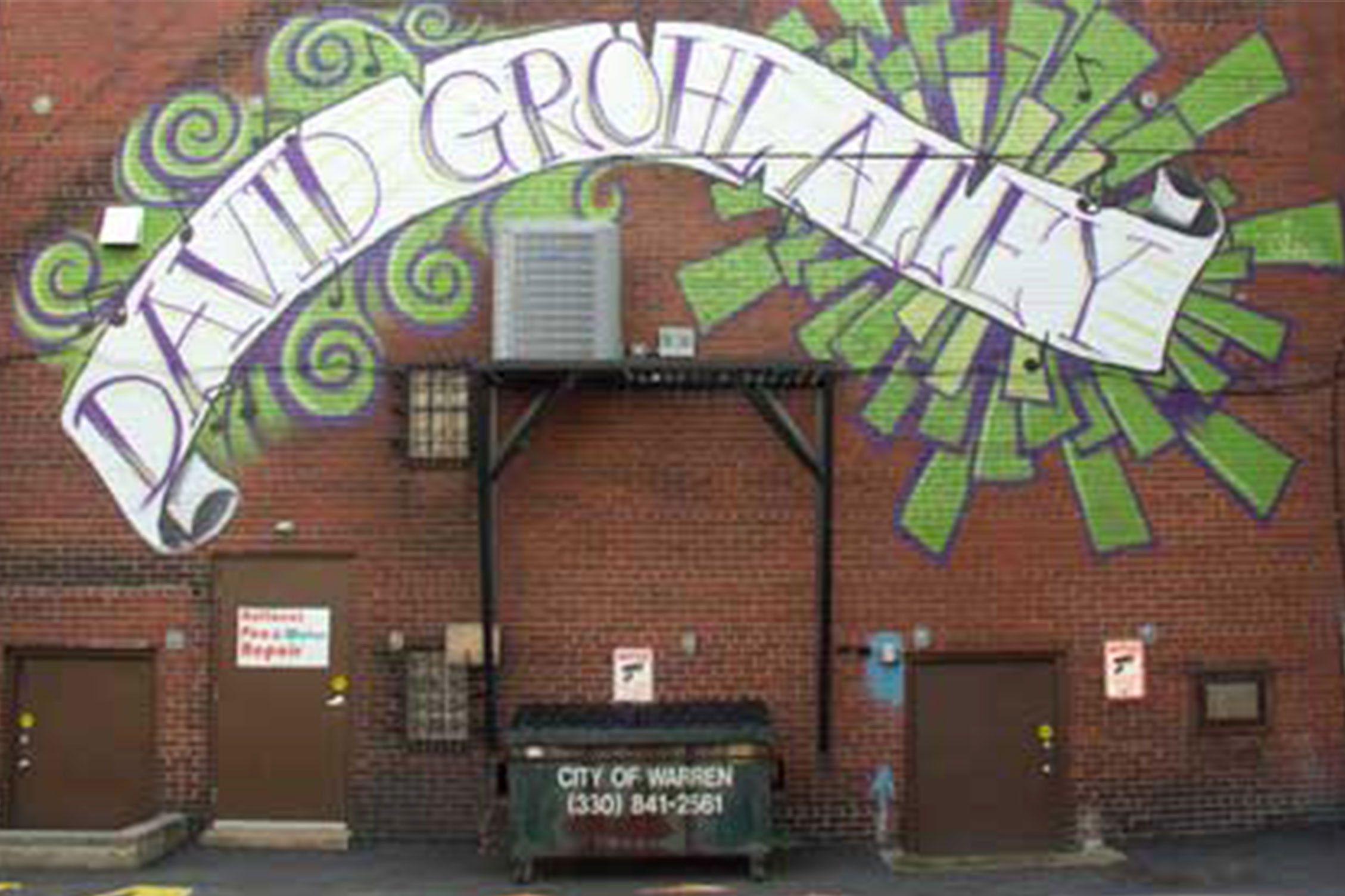 Graffiti mural on a brick wall displays "DAVID GROHL ALLEY" in vibrant swirls, with a nearby dumpster labeled "City of Warren" in an alleyway.