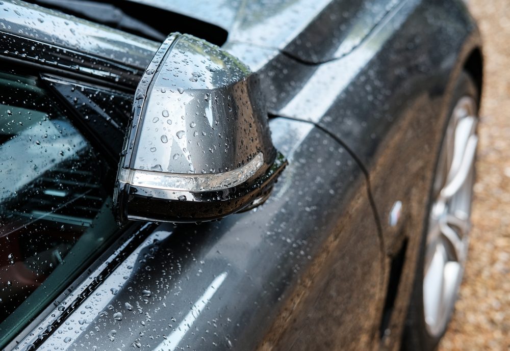 A car side mirror is covered with raindrops, attached to a sleek, dark, wet vehicle exterior with a visible tire and gravel ground.