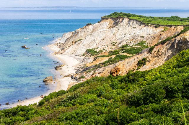 Cliffs rise steeply above a sandy beach, bordered by lush greenery, with calm turquoise ocean extending to a distant horizon.
