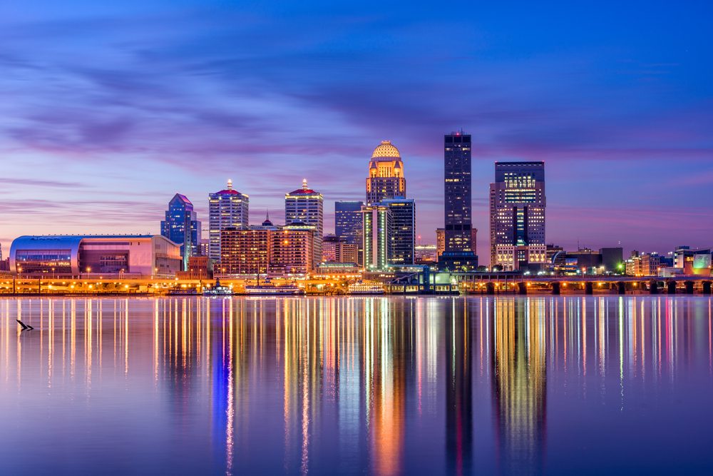City skyline reflects vibrantly on calm water, with multicolored lights under a twilight sky, depicting an urban waterfront at dusk.