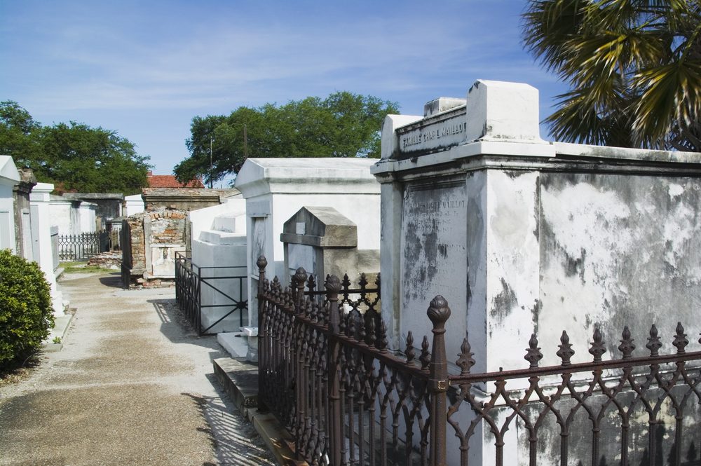 Tombs stand in an old cemetery, textured with age, surrounded by wrought iron fences under a bright blue sky and various green trees.