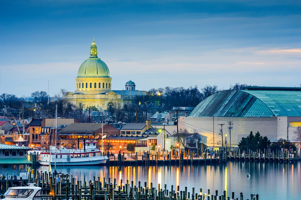 Dome-topped building illuminated at dusk, reflecting on water, surrounded by smaller buildings and a harbor with boats, under a blue sky.