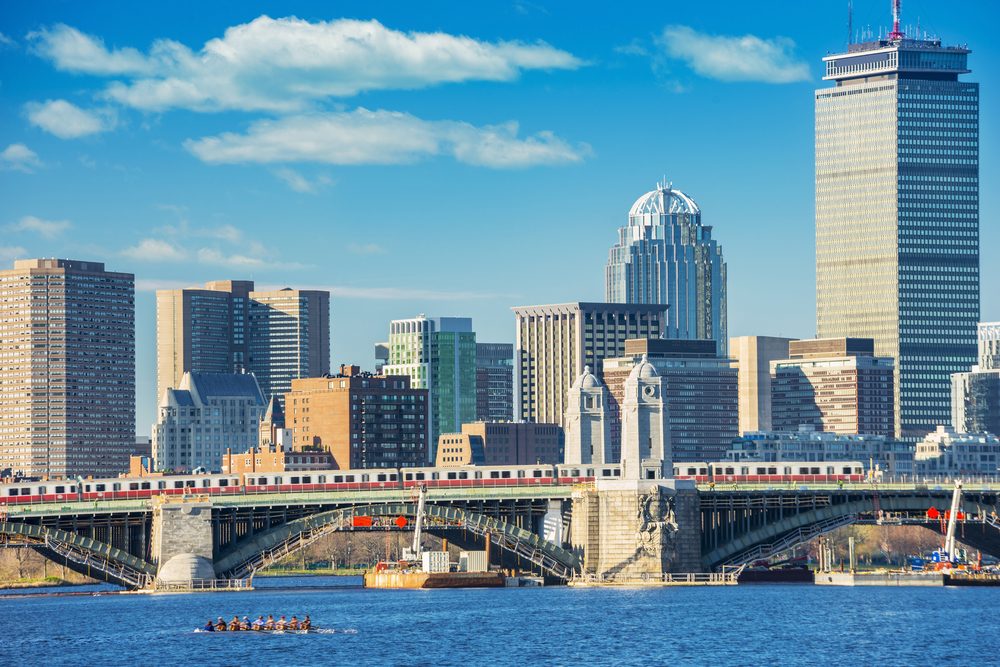 Rowboat glides across water, beneath a bridge, with a city skyline and a train on the bridge in the background under a blue sky.