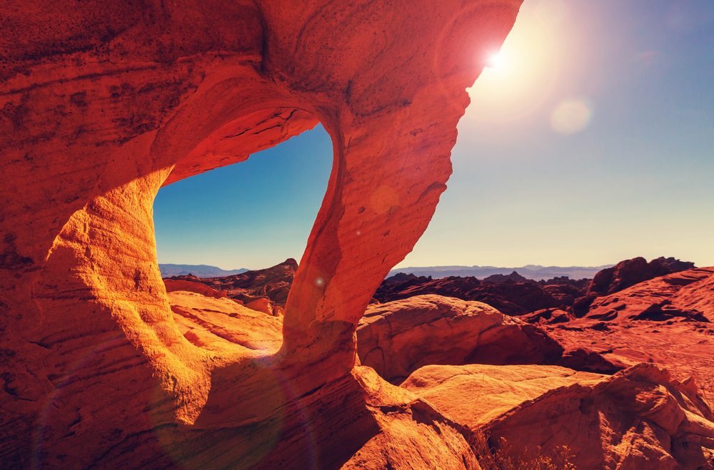 Rock formation frames sunlit desert landscape with clear blue skies and distant mountains, creating a natural window effect.