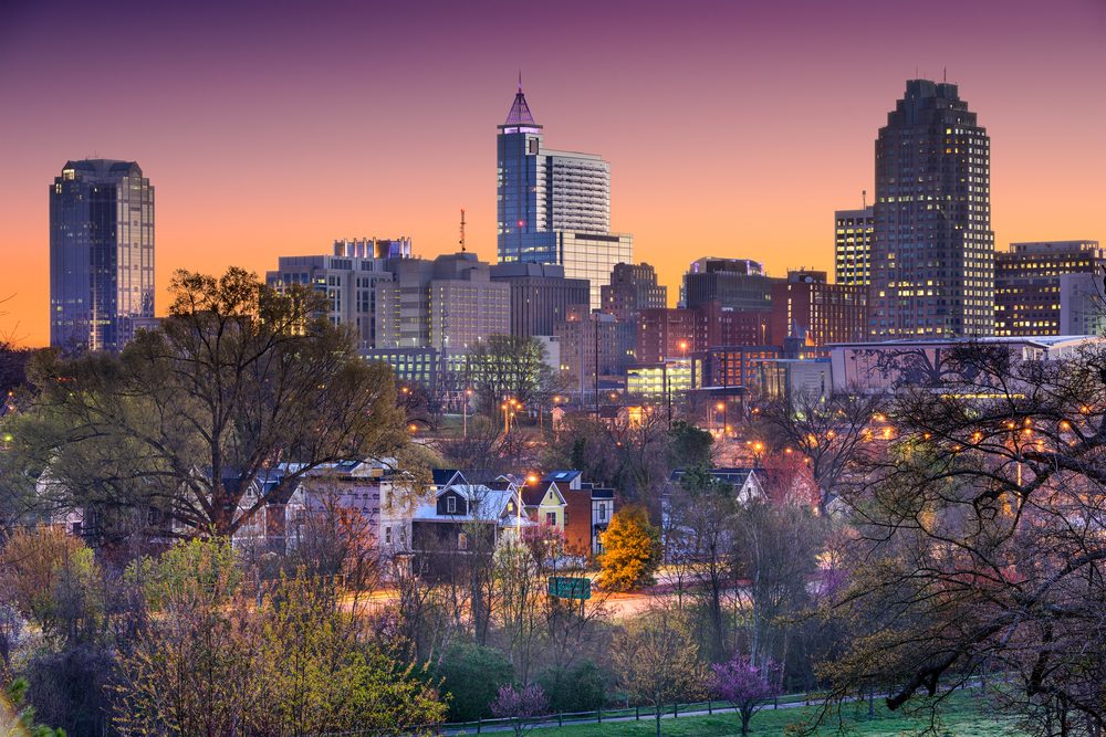 Buildings form a skyline against a golden sunset, surrounded by trees and city lights, providing a contrast of urban and natural scenery.