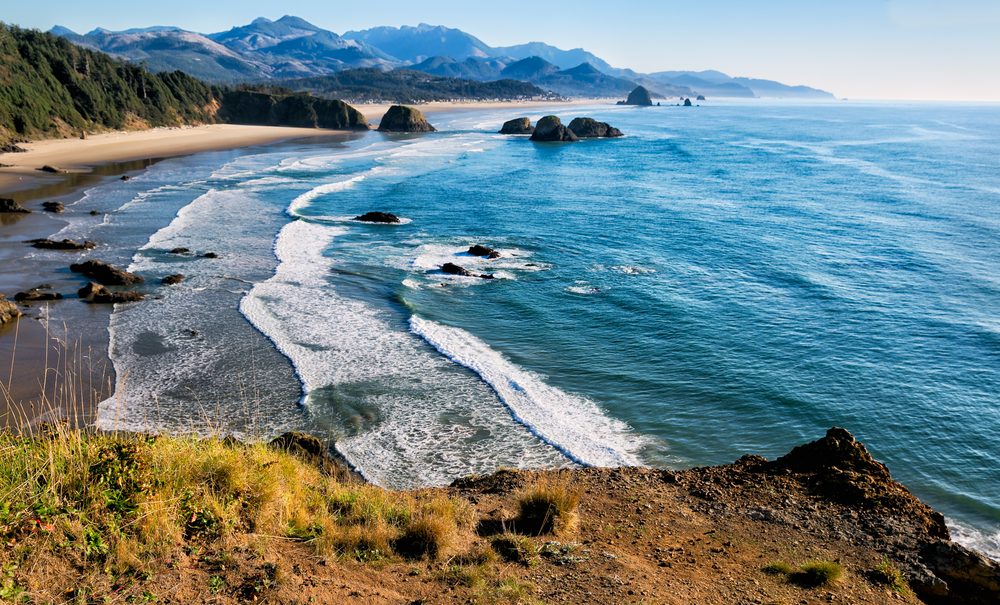 Waves gently crash on sandy beach, bordered by rugged cliffs and dense forest, with distant mountains under clear blue sky.