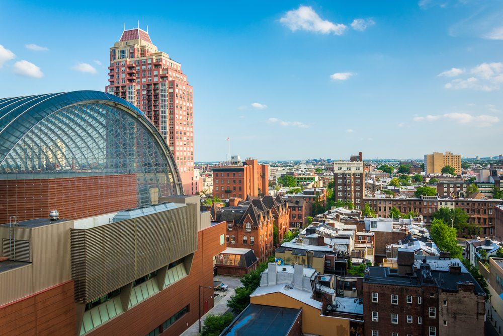 Large glass-domed building stands beside residential structures under a clear blue sky, offering an urban cityscape view with scattered trees and distant skyline.