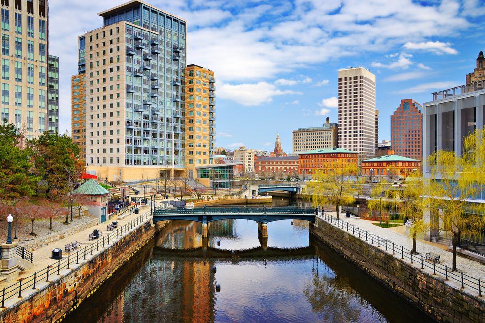 Tall buildings overlook a calm river, reflecting the cityscape, with a bridge crossing over, framed by trees and a clear blue sky.