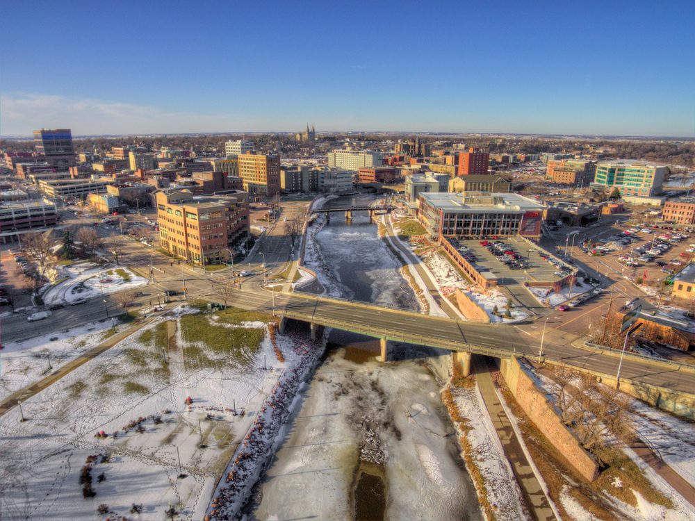 Aerial view of a city with buildings; bridge crosses a snowy river; streets and parking lots are visible under a clear blue sky.