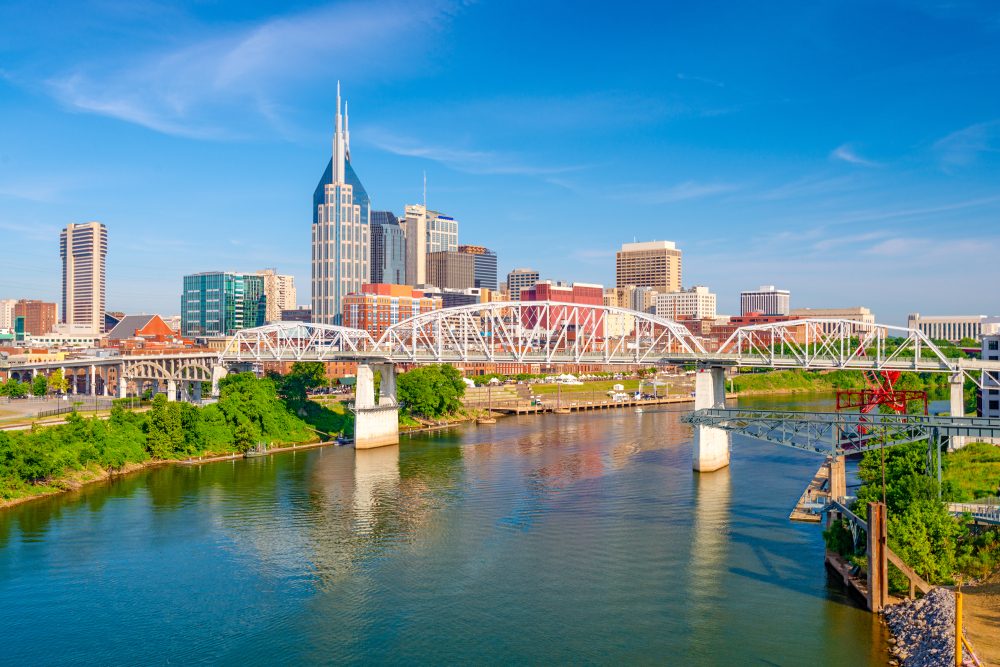 A white bridge crosses over a river, with a cityscape of tall buildings and a clear blue sky in the background.