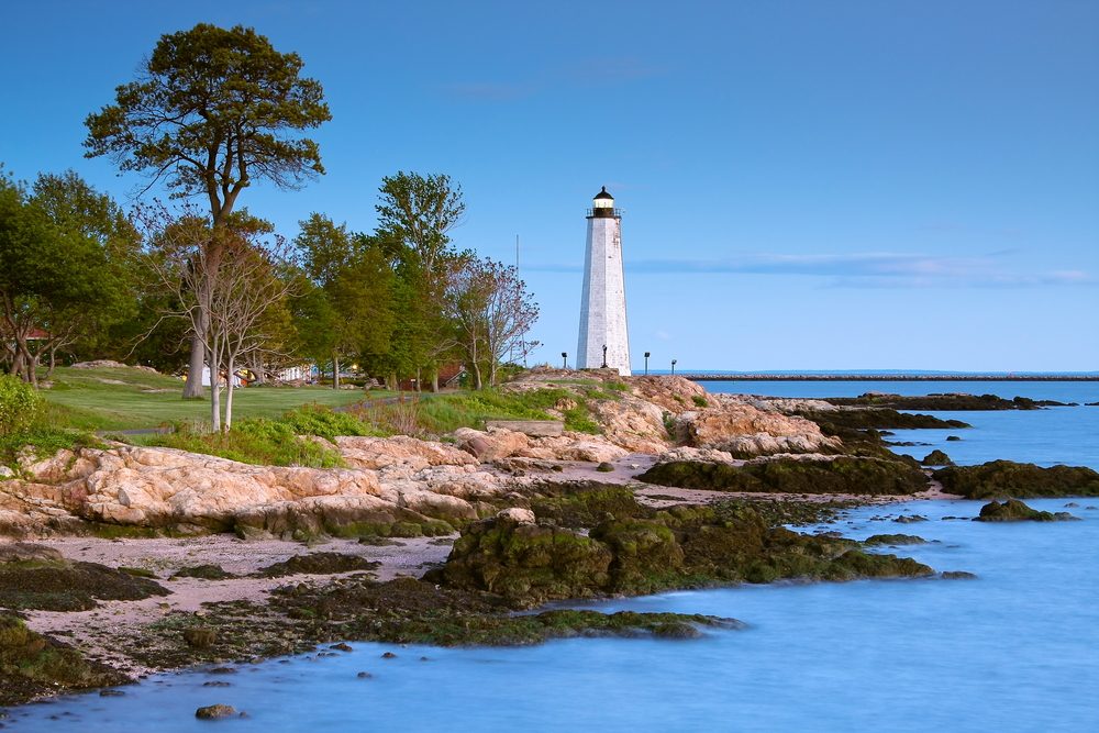 Lighthouse stands tall on rocky shoreline, surrounded by green trees and calm blue ocean under a clear sky.