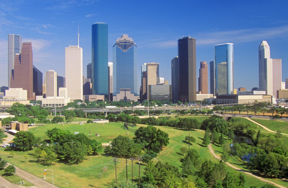 Skyscrapers stand tall under a clear sky, overlooking a green park with a winding path in a bustling urban landscape.