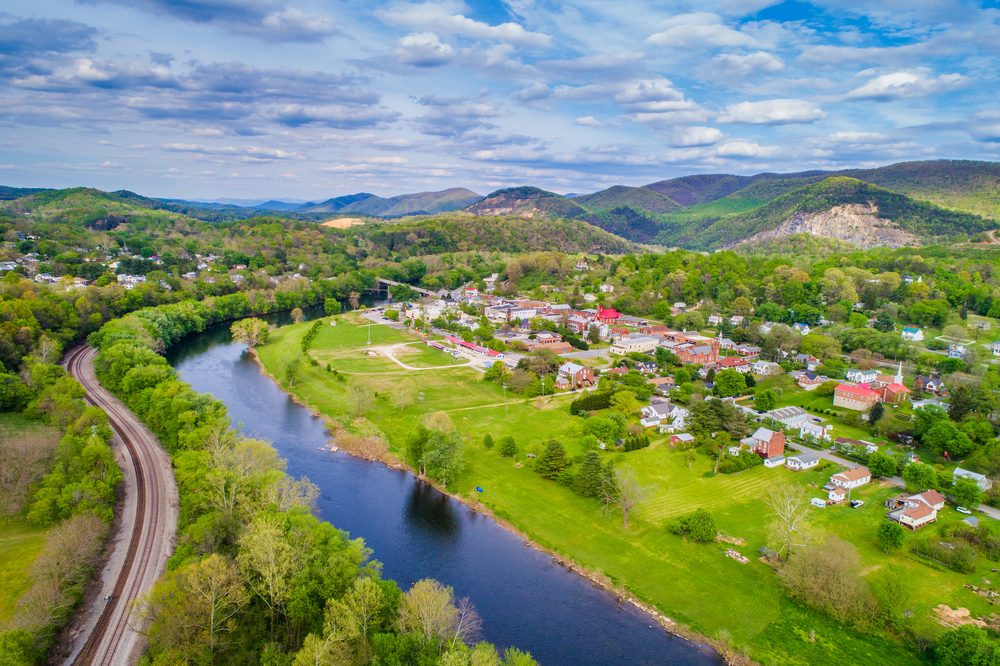 River curves through lush green landscape, bordered by railway tracks, with a small town nestled against mountainous backdrop under a partly cloudy sky.