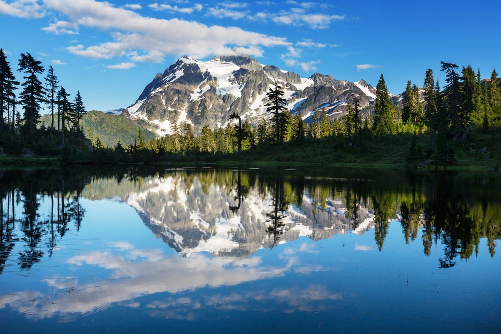 Mountain reflected in a calm lake, surrounded by evergreen trees under a clear blue sky with scattered clouds.